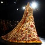 A glamorous slice of pizza styled as a fashion icon, adorned with gold leaf and glitter, walking a runway under bright lights with a cheering audience.